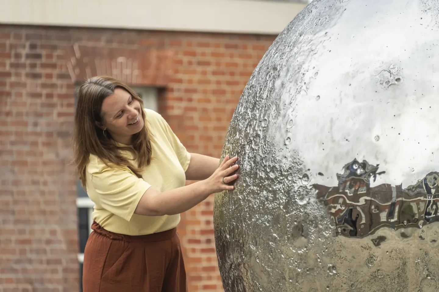 A smiling woman in a yellow t-shirt runs her hands over the craters of Luke Jerram's silvery Mirror Moon sculpture at the Royal Observatory in Greenwich