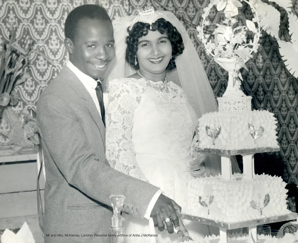 Black and white photograph of a couple on their wedding day. The man is on the left, wearing a suit and black tie, while the woman is on the right in a white wedding dress with lace detailing on the arm. They are standing next to a multi-tiered wedding cake