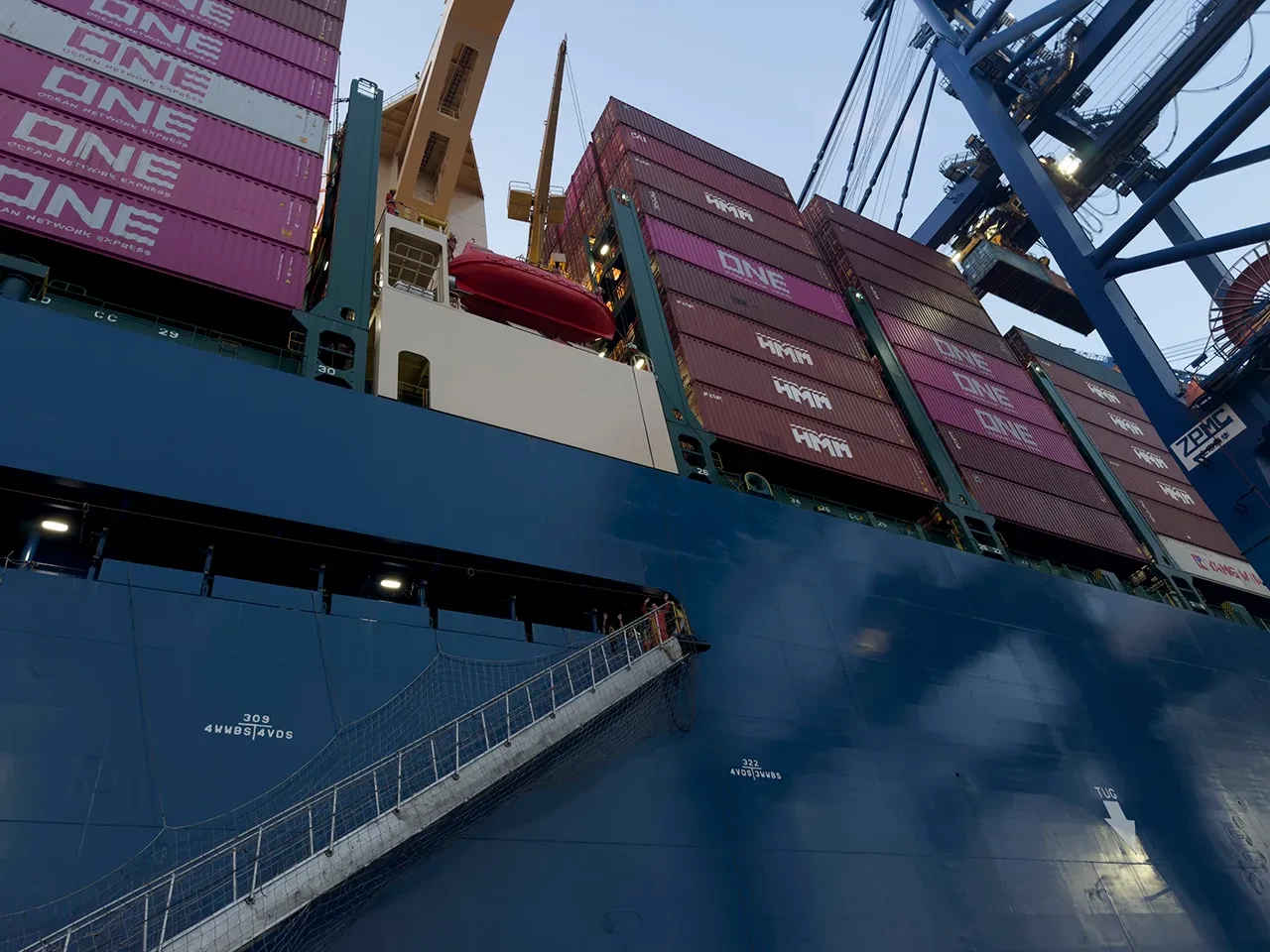 View from the quayside looking up a a huge container ship. Crew members are just visible waving from the top of a ladder that leads up to the deck