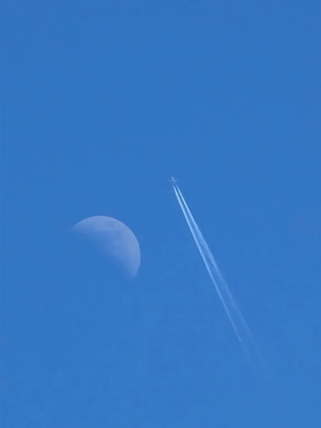Daytime photo against blue sky showing partial moon with plane trail next to it