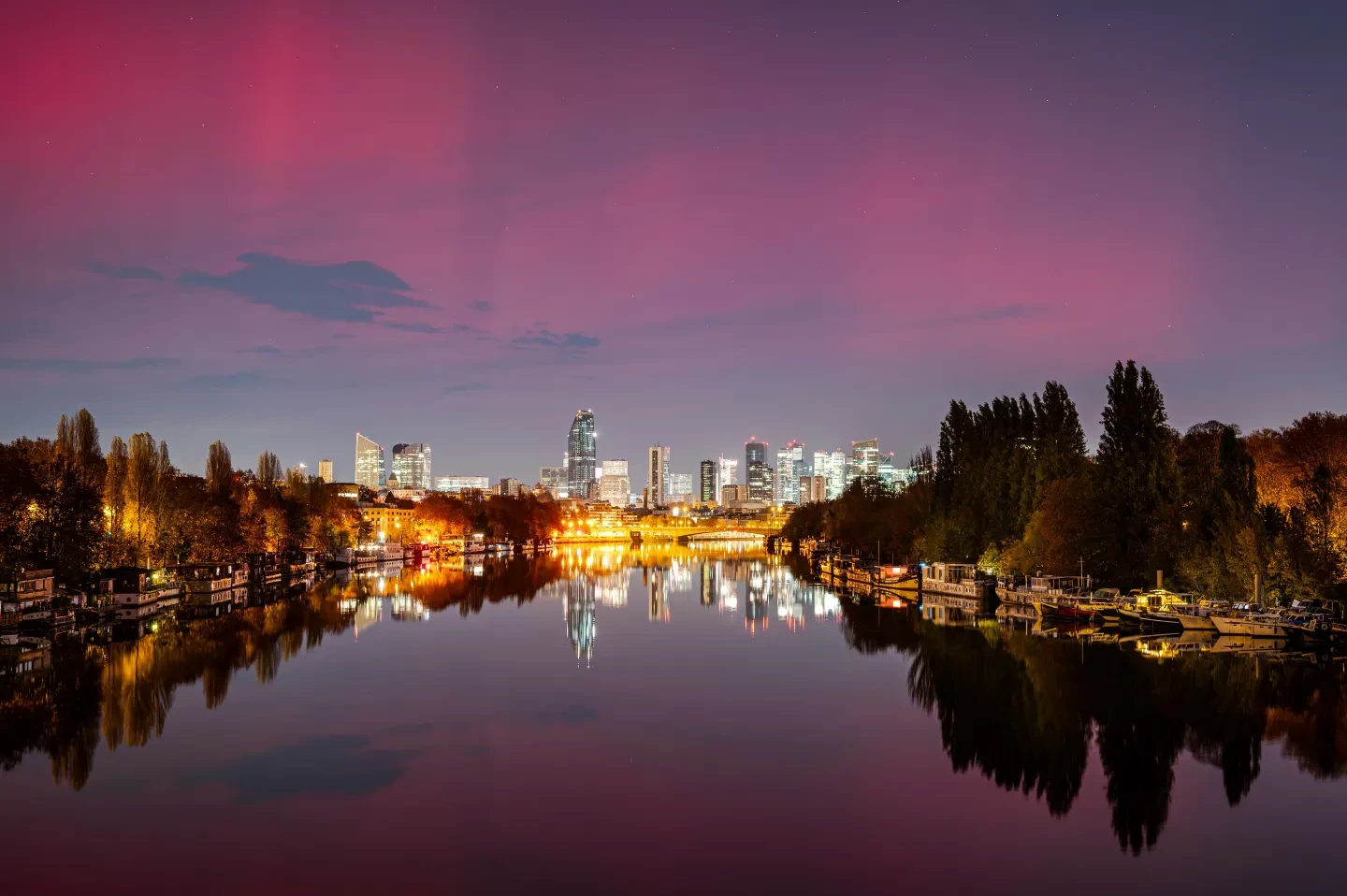 A tree-lined river in the foreground gives way to a light-speckled cityscape behind, with a pink aurorae-smudged sky 