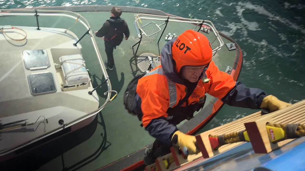 A harbour pilot climbs a ladder to board a ship. He is wearing orange overalls and orange helmet, and the boat he has just stepped off from is visible below