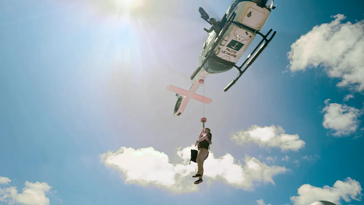 A man descends from a hovering helicopter by hoist. He is harnessed and holding on to a rope attached to the helicopter, while a person in the aircraft leans out to watch his progress