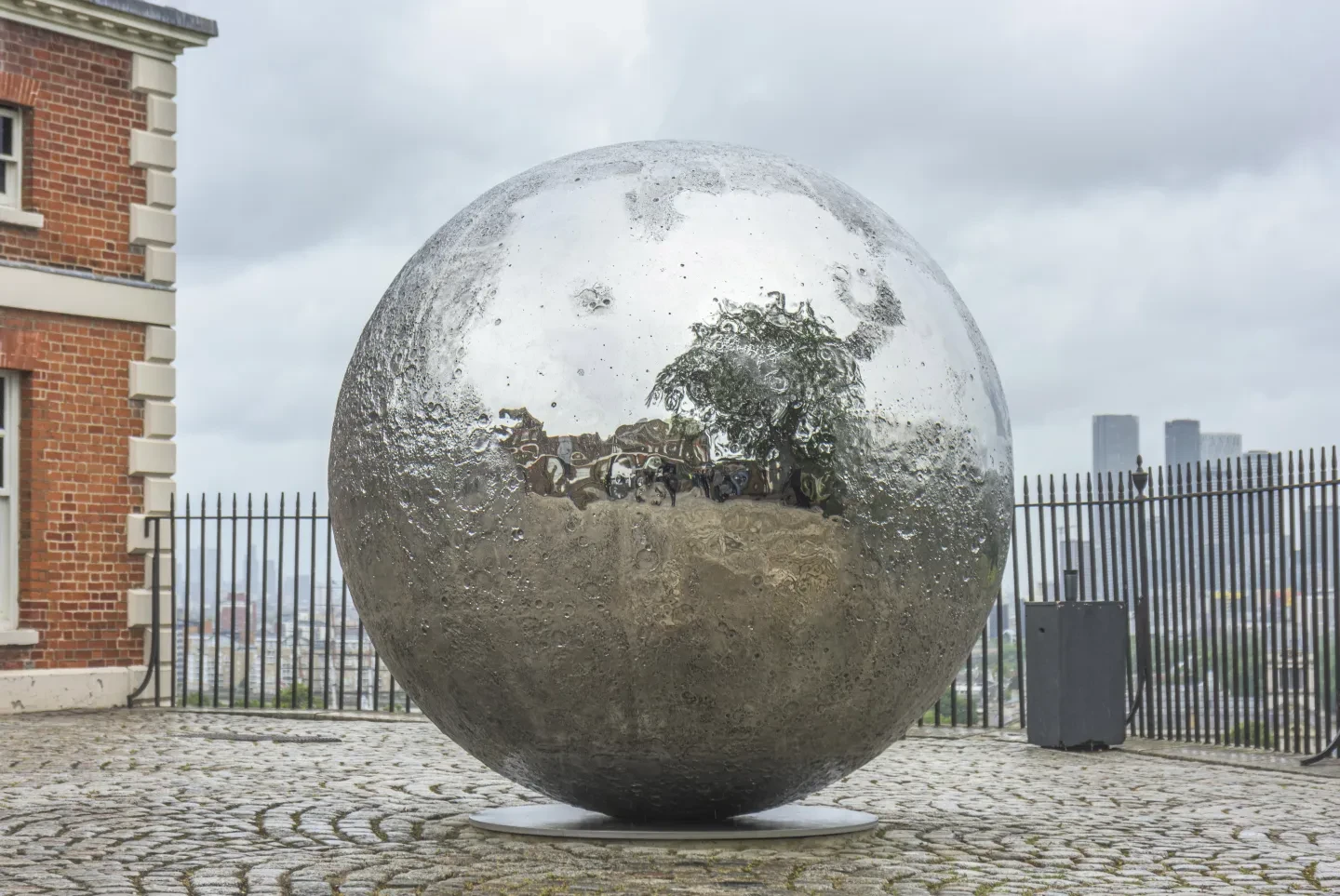 Large mirrored moon sculpture in Royal Observatory courtyard with city skyline in background