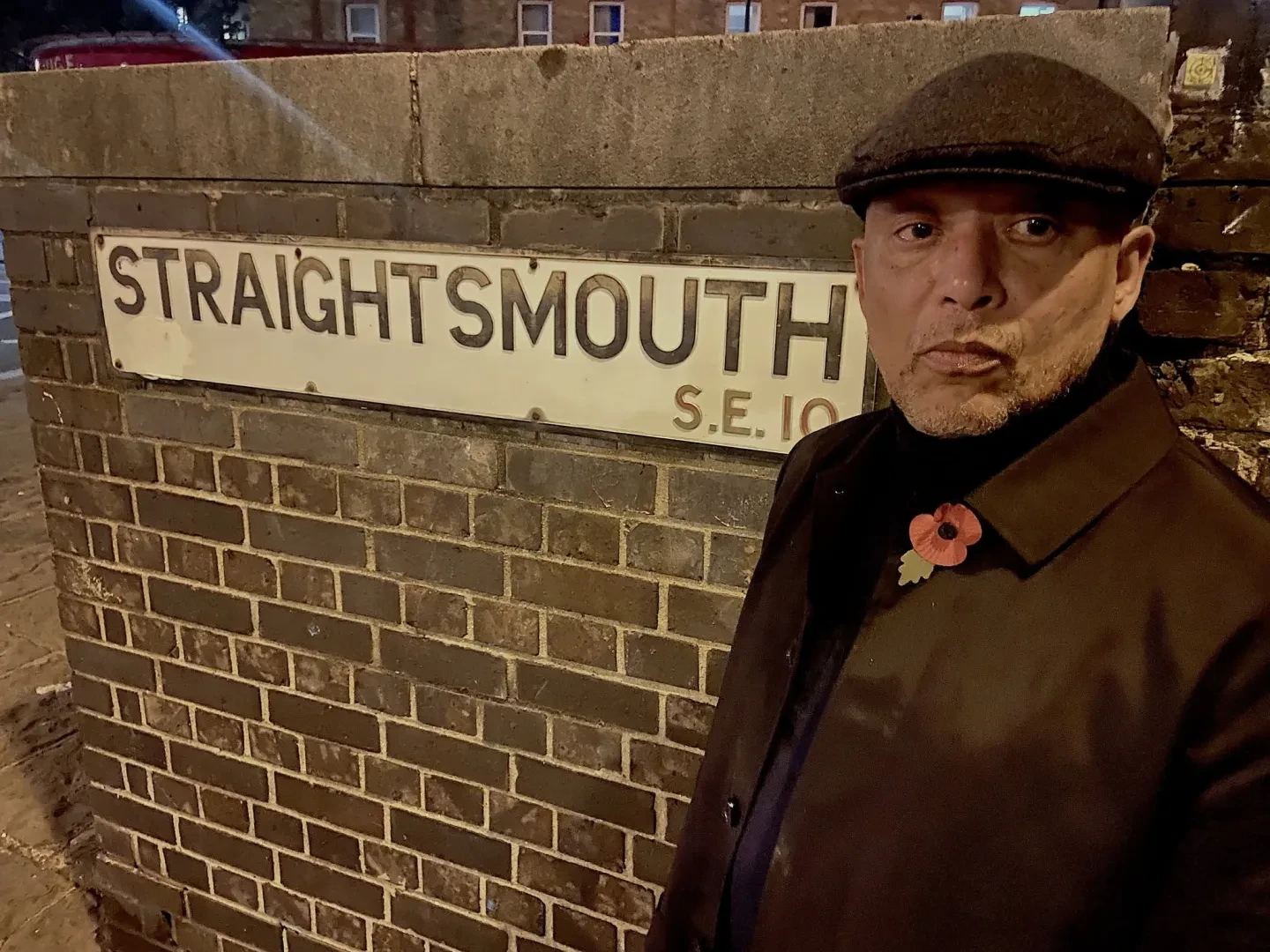 A colour photo with a man standing against a wall to the right of the photo. And a sign on the wall that says Straight Mouth SE10. The man is wearing a brown cap and coat with a poppy badge on the lapel