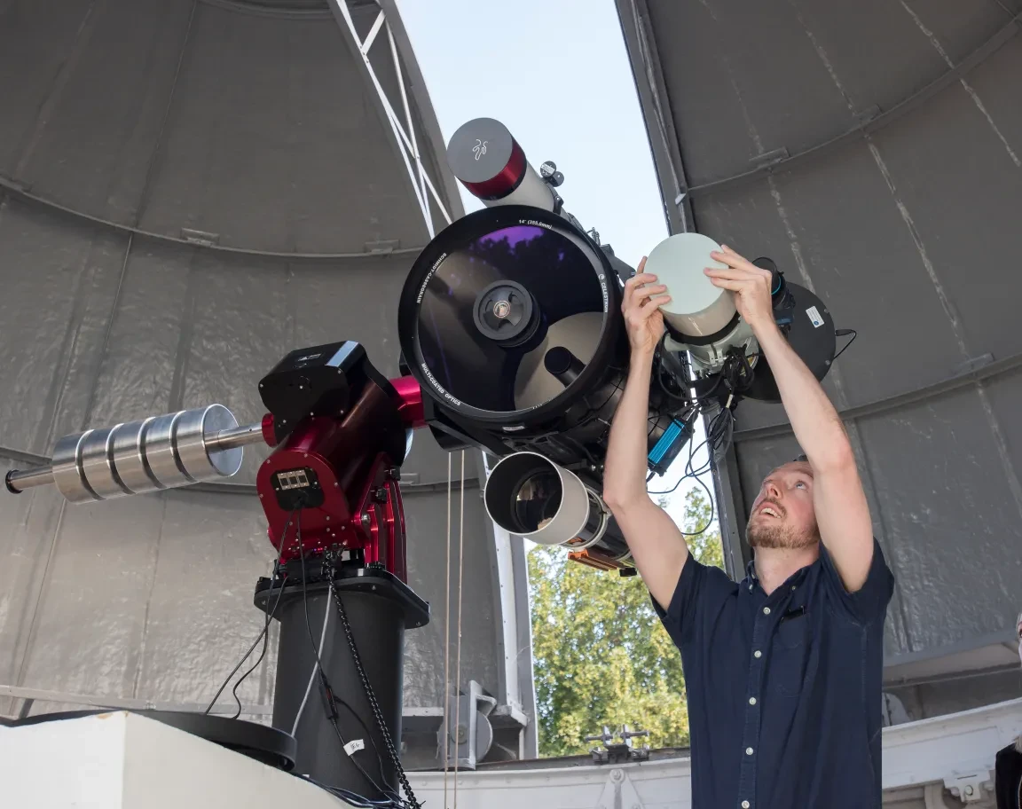 A man adjusts a large telescope under a partially open domed roof.