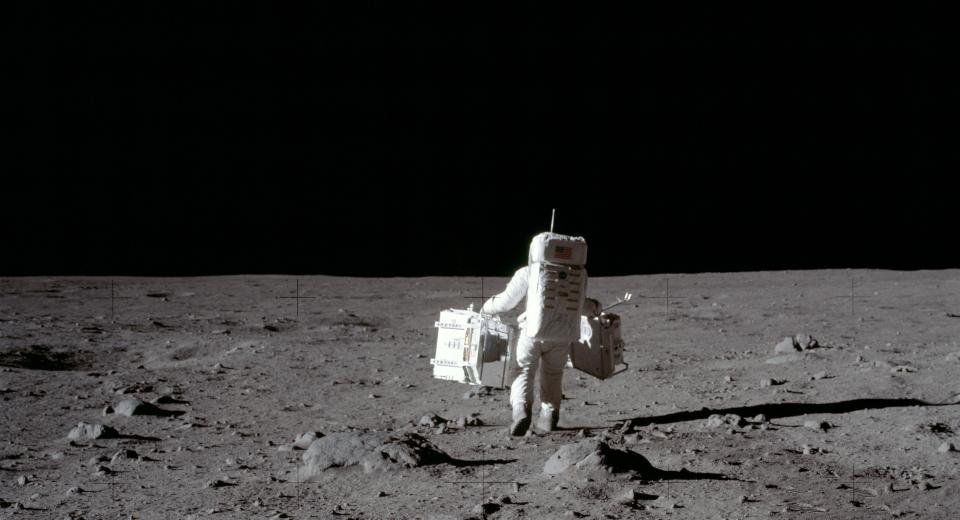Apollo era astronaut walking away across the surface of the Moon, carrying equipment in both hands. 