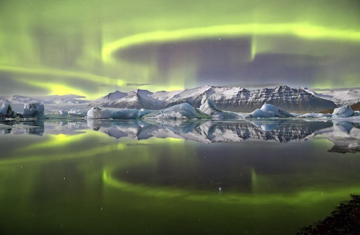 A ring of neon green aurora is reflected in a lagoon, surrounded by glaciers.