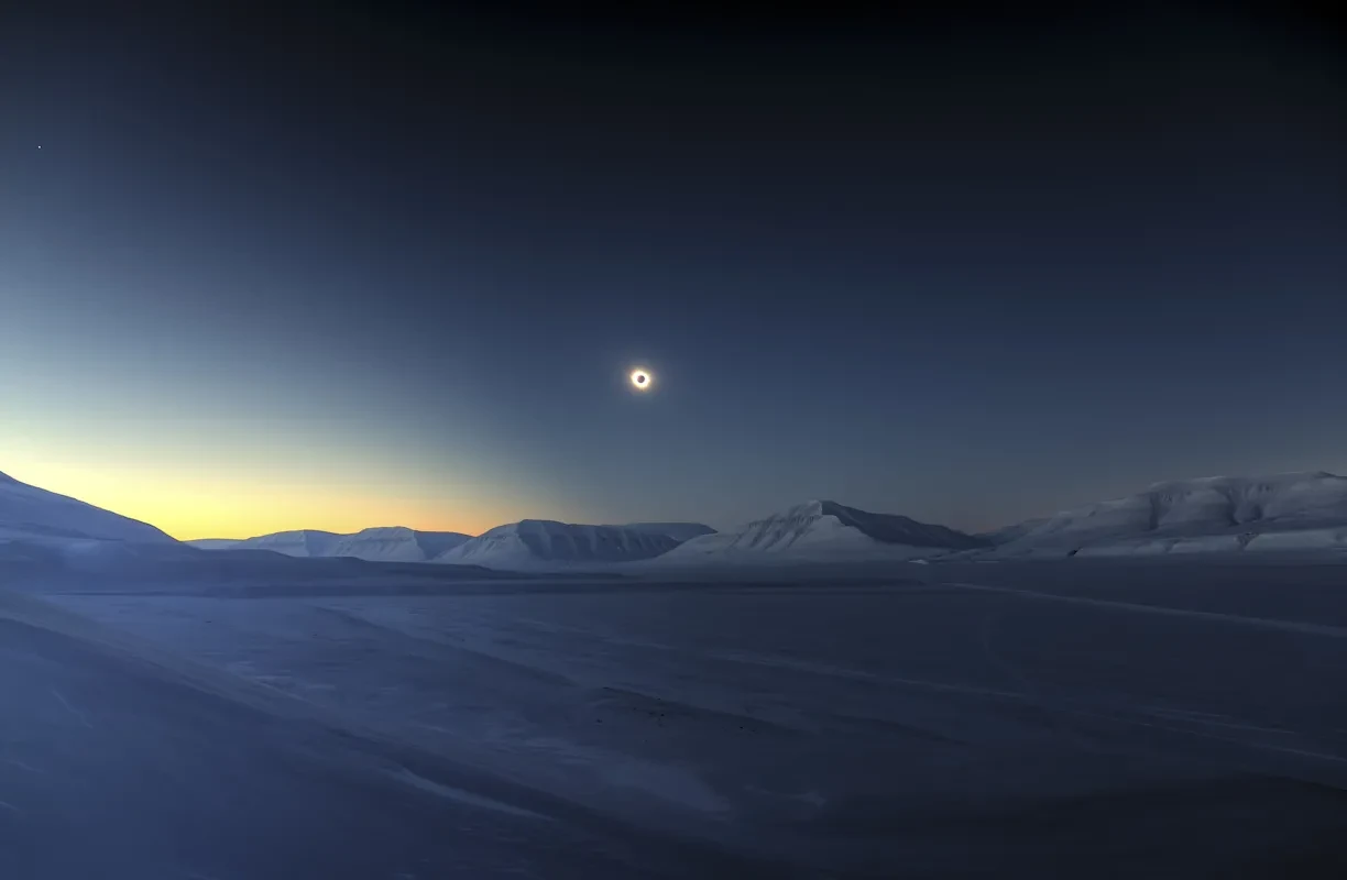 A solar eclipse in the dark sky above snowy plains and mountains.