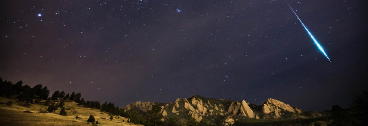 photograph of the night sky over a countryside landscape. One bright meteor visible in the sky. 