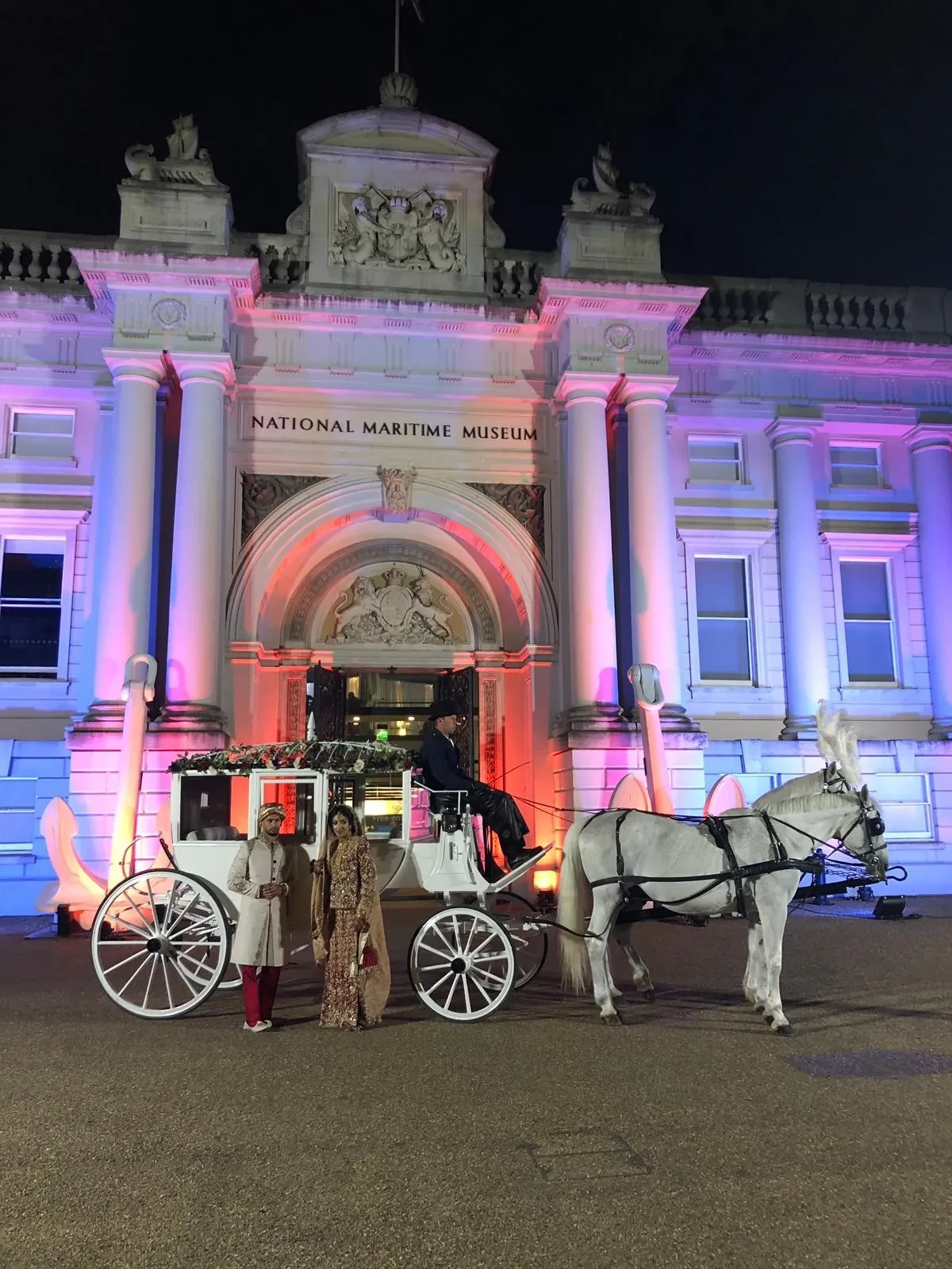 A couple in Hindu wedding attire with a white horse-drawn carriage outside the National Maritime Museum.