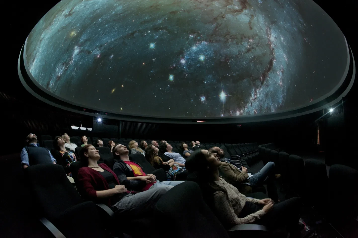 An audience in reclined chairs, looking up at a planetarium show on a domed ceiling.