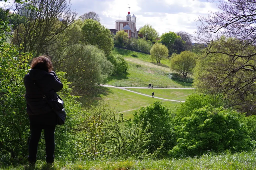 A person stands in lush greenery in Greenwich Park, taking a photo of Flamsteed House in the distance.