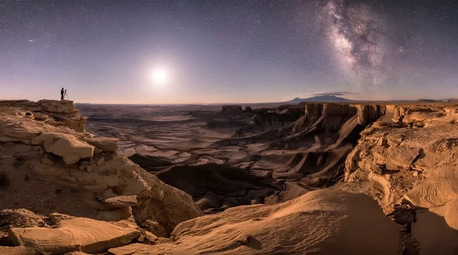 A galaxy is visible in a starry sky above a distant photographer in a dramatic canyon landscape.