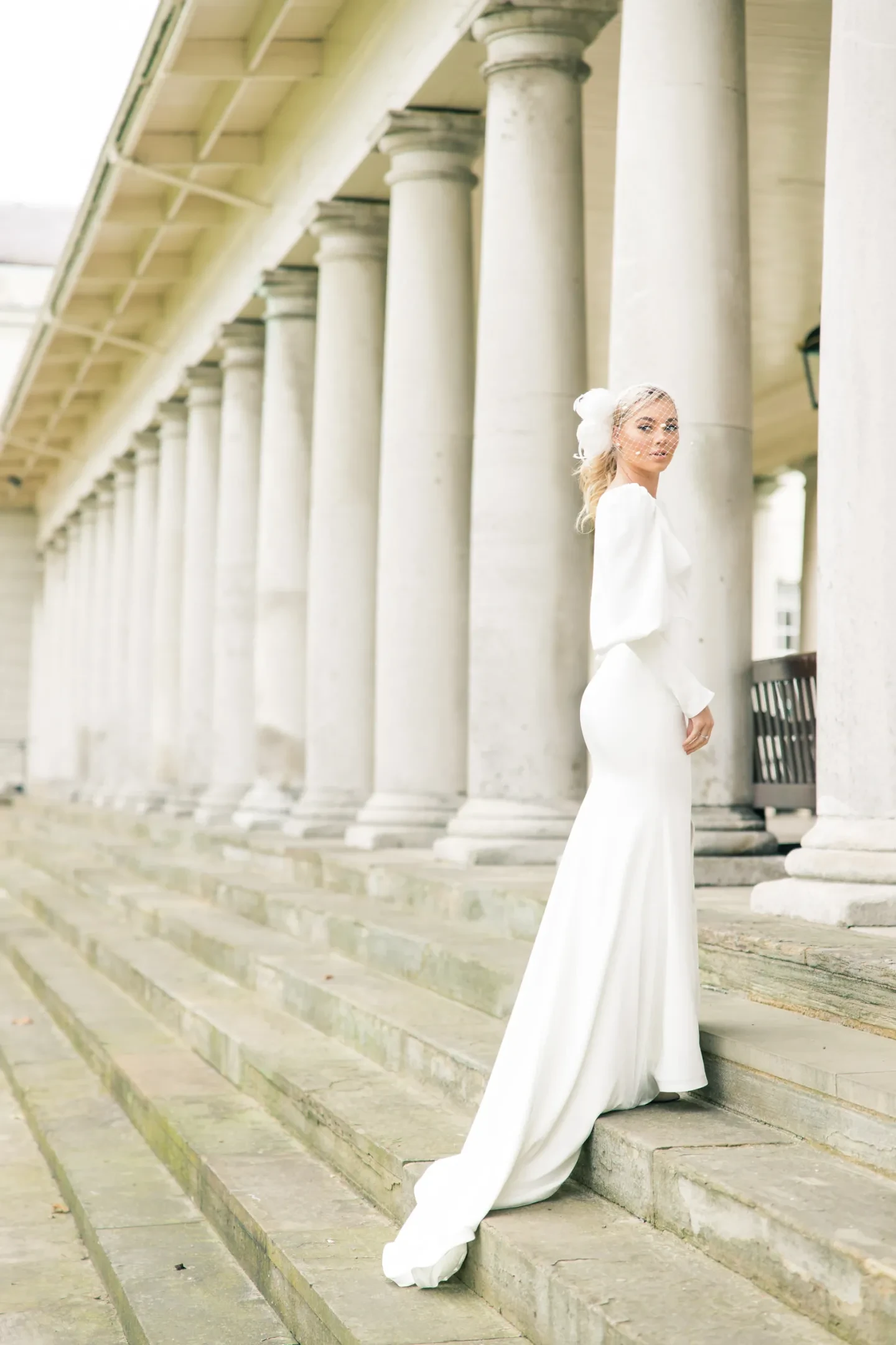 A bride in white veil and long dress on steps next to a row of white columns.