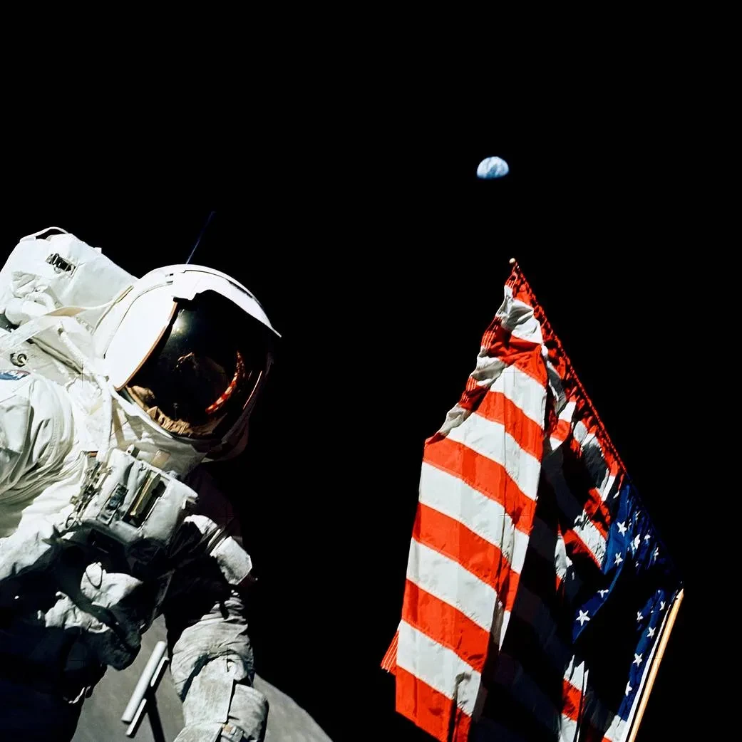 An astronaut in their space suit stands next to an American flag against a black sky.