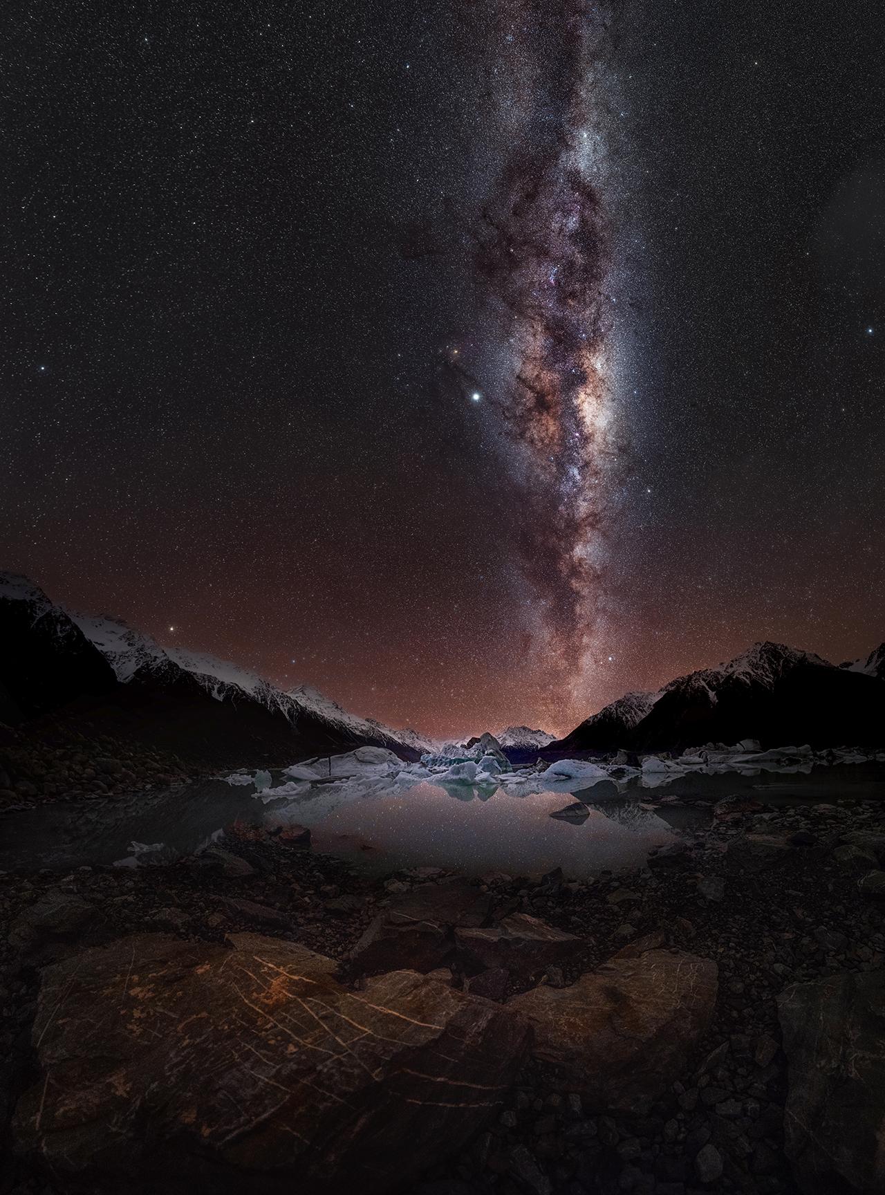The Milky Way in tones of violets and purples and browns in the sky behind snowy mountains and a lake