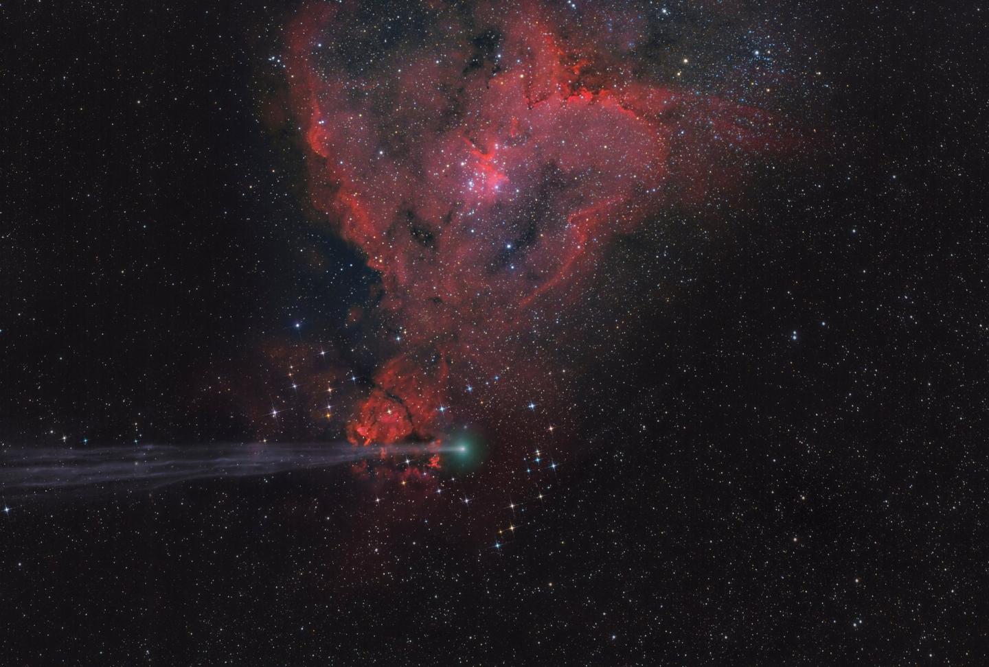 A photograph of a heart shaped emission nebula, in red, with a comet streaking across in the foreground. 
