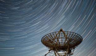Image of radio telescope in the foreground and in the night sky behind it are semicircular star trails 