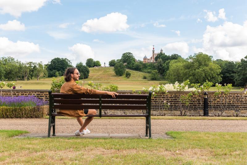 A man sits on a park bench beside Greenwich Park, with the Royal Observatory in the background
