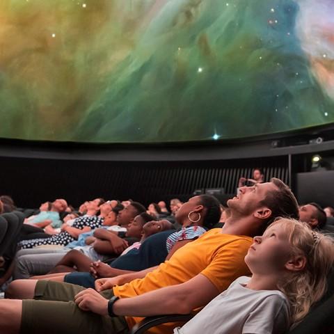 A large group of people, watching a planetarium show. The screen is showing a swirl of a galaxy