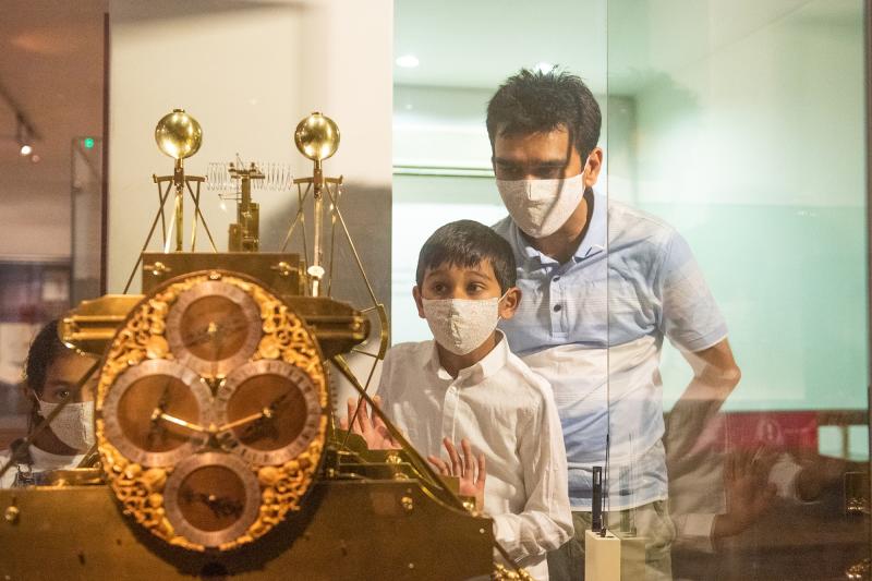 A father and son, both wearing masks, look at the display of the Harrison Clocks inside the Royal Observatory