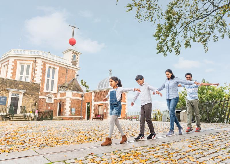 A family plays on the Prime Meridian Line in front of the historic buildings of the Royal Observatory