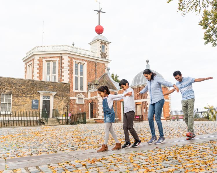 A family plays on the Prime Meridian Line in front of the historic buildings at the Royal Observatory Greenwich