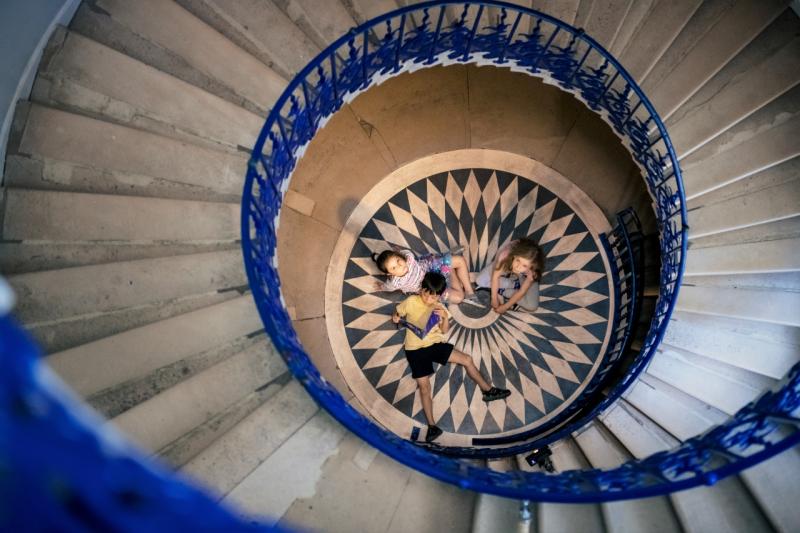 A group of children sit on the floor, looking up at a spiral staircase