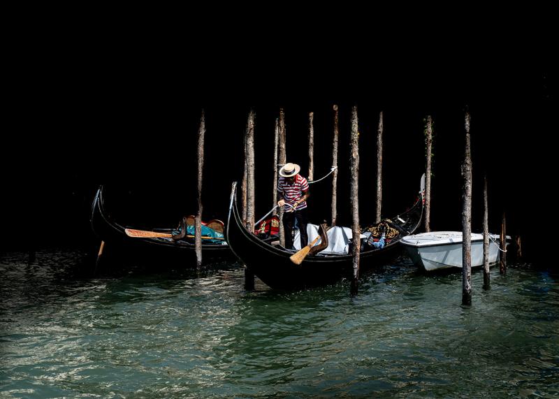 A gondolier ties up a gondola at a mooring poll in Venice. He is caught in the sunlight, but the rest of the photo is in shadow