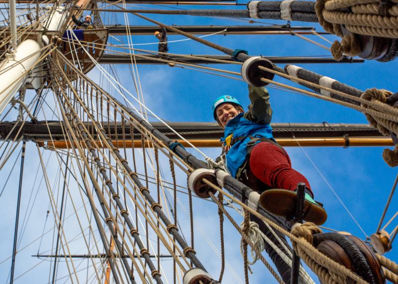 A woman climbs the rigging of historic sailing ship Cutty Sark. She is looking down at the photographer on deck, and the rigging and masts are above her