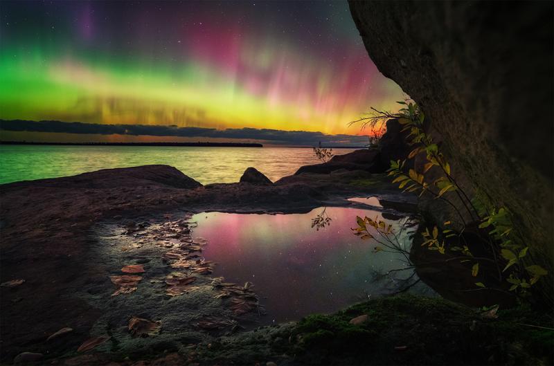A landscape photograph at night showing the aurora in beautiful green and pink, reflected in a calm sea