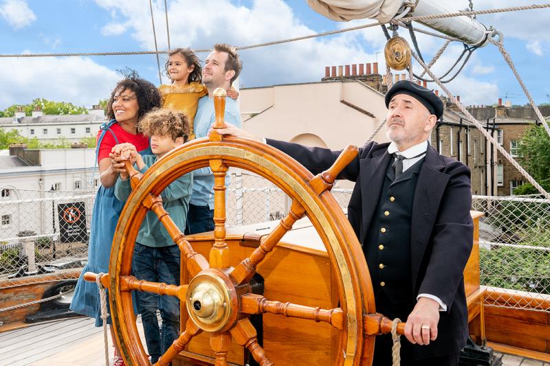 A smiling family of four look out on the main deck of Cutty Sark, with a character actor dressed as the ship's captain holding the ship's wheel