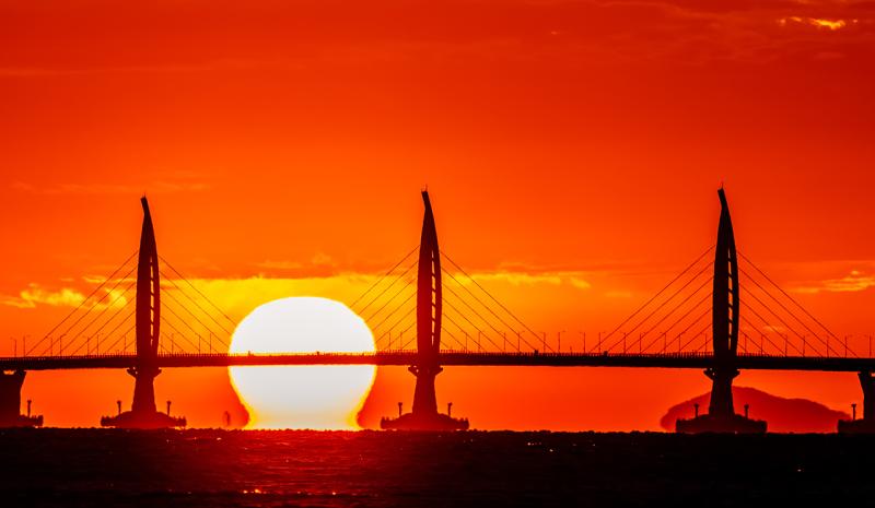 Image of a silhouette of a bridge in Hong Kong with orange and yellow sunset sky behind it, the Sun appears to be sitting on the water and the bottom of it is distorted in a mirage