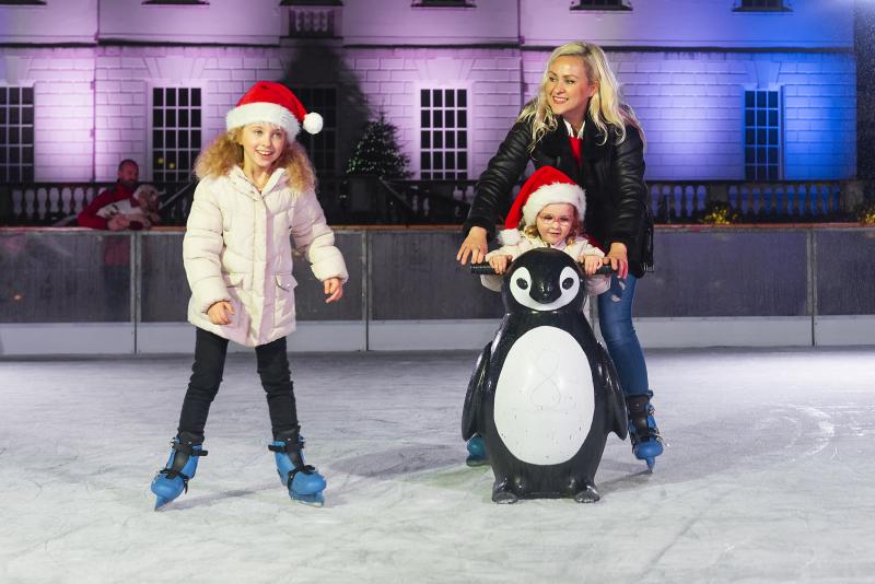 Image of ice rink at night time with house lit up in purple and blue lights behind three people, a mother with her two children, one is younger and using a penguin skate aid