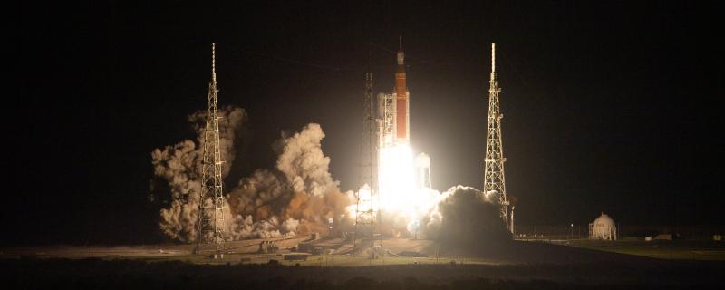 Image of orange rocket at night with white-hot flames coming out of the bottom and propelling it off the ground, a trail of grey smoke at the bottom