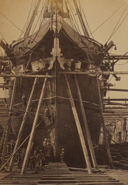 A sepia tone photograph showing a ship in a dry dock. Wooden struts support the hull, and men are visible at the front of the ship working on the bow