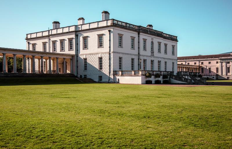 A view of the Queen's House in Greenwich from the outside. The square white building is seen from an angle, with green grass stretching up to the main entrance