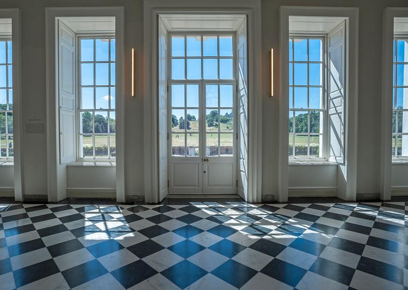 Photograph looking out through the tall windows of the part of the Queen's House known as the Orangery. The floor is designed in a black and white marble checkerboard pattern, and the windows allow a beautiful view across Greenwich Park