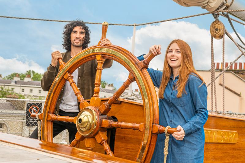 A couple pose holding the ship's wheel of Cutty Sark, the historic ship and visitor attraction in Greenwich