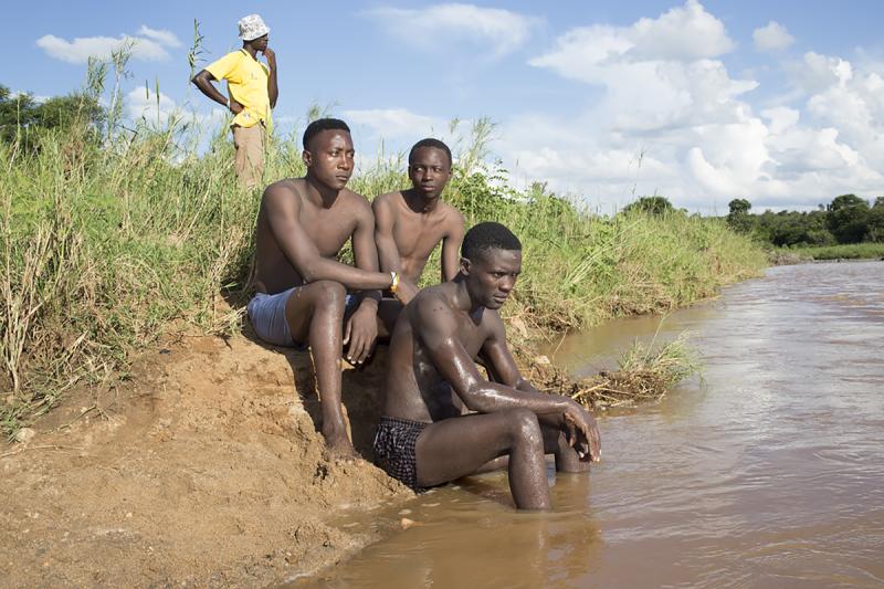 A group of three Black men in swim shorts sit on the muddy bank of a river or lake. Another man in the background in a yellow shirt and white hat looks out across the water