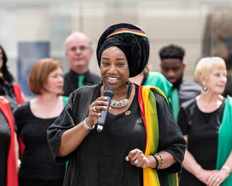 A female singer performs a solo as part of a choir performance. She is holding a microphone and is wearing a black dress with a multicoloured sash over her left arm, with a matching headscarf