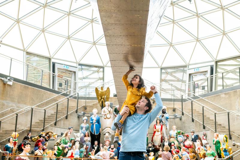 A father lifts his daughter up to touch the hull of historic ship Cutty Sark. A colourful display of ships' figureheads is arrayed in the background