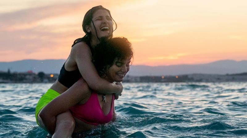 A promotional picture for the Netflix documentary The Swimmers. Two women in bathing costumes are in the water at sunset, one on the other's shoulders. They are both smiling