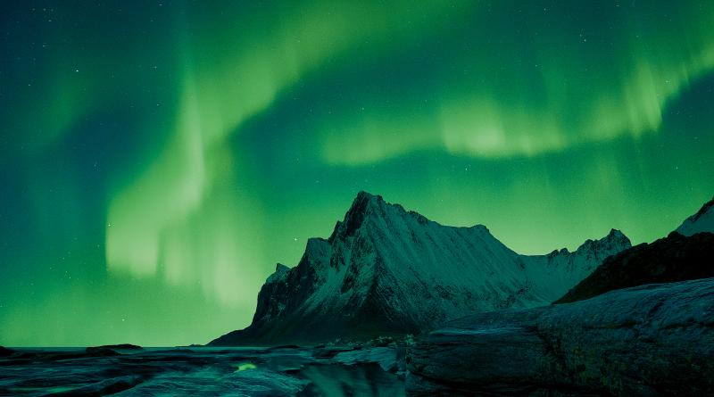 Image showing mountain and rocks in foreground, with rippling emerald aurora in the sky behind the mountain, which is reflected in a pool of water