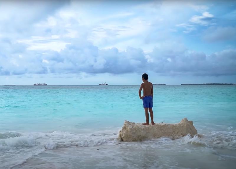 A boy stands on a rock looking out to sea. The waves are lapping around him