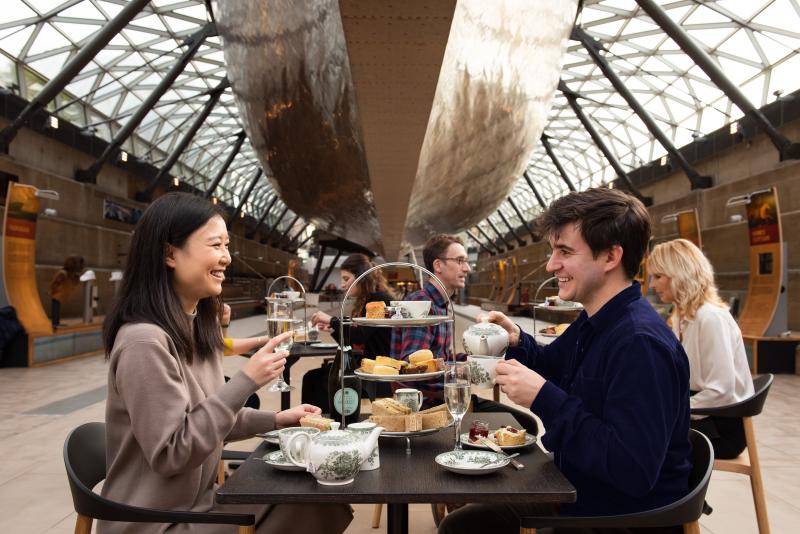 A couple enjoy afternoon tea beneath historic ship Cutty Sark. On the table are cakes, sandwiches and fine china. Above them, the hull of the historic ship can be seen, almost 'floating' thanks to the steel support structure and glass roof