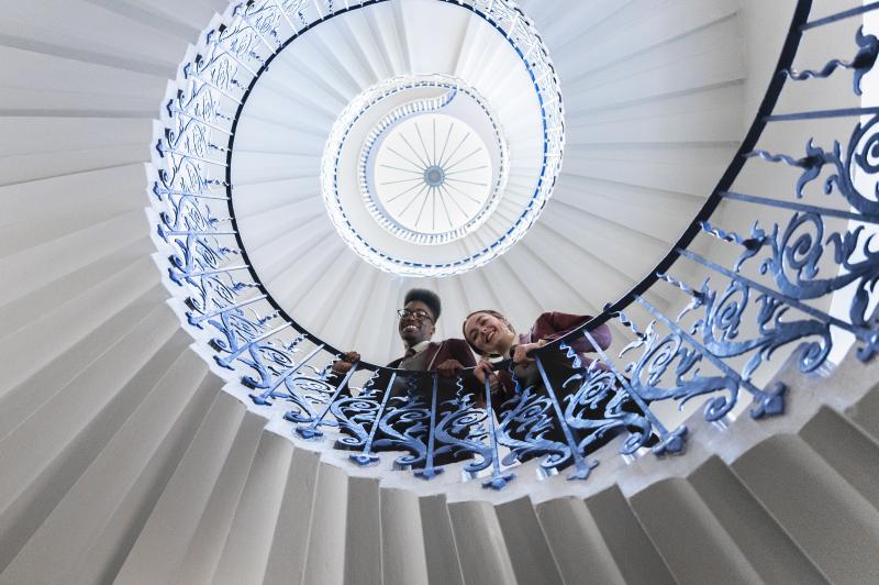 Two students in dark red blazers look over the bannisters of the Tulip Stairs in the Queen's House Greenwich