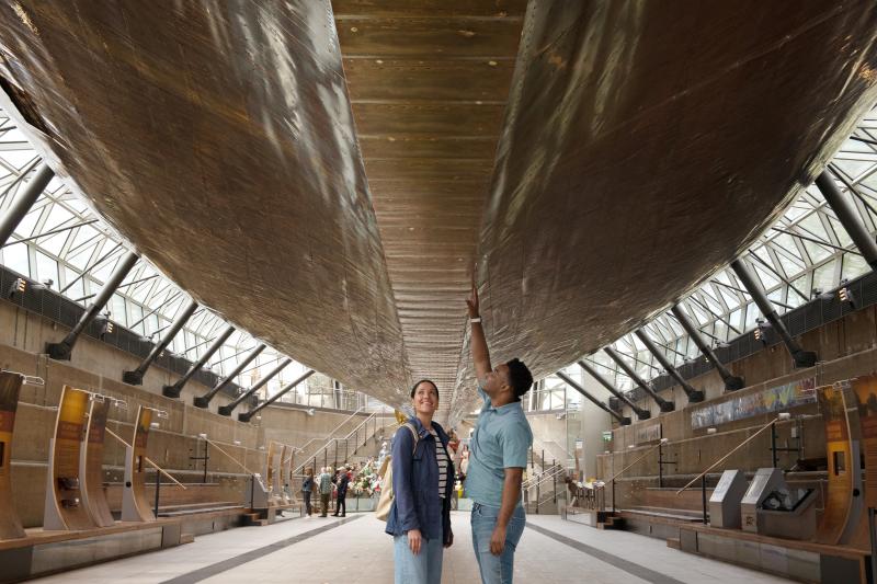 A couple look up at the hull of Cutty Sark. The ship appears to be floating above them, supported as it is by an array of steel beams