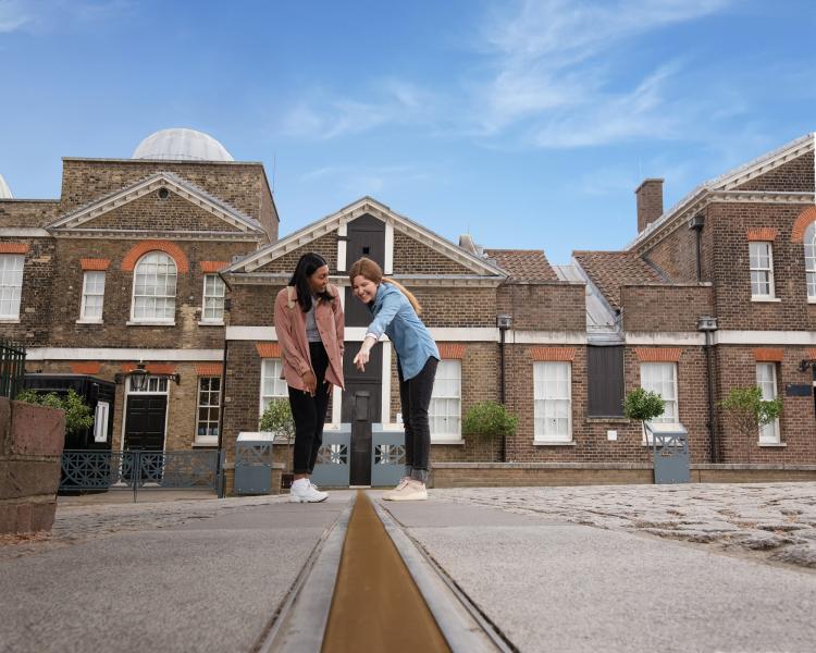 Two friends stand and point at the Prime Meridian Line in the courtyard of the Royal Observatory Greenwich